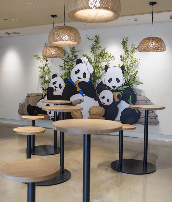 An indoor seating area with wooden tables and hanging lights is set against a large mural of the Bao Family pandas holding cookies and bamboo. The warm, inviting atmosphere is themed for the panda birthday celebration.