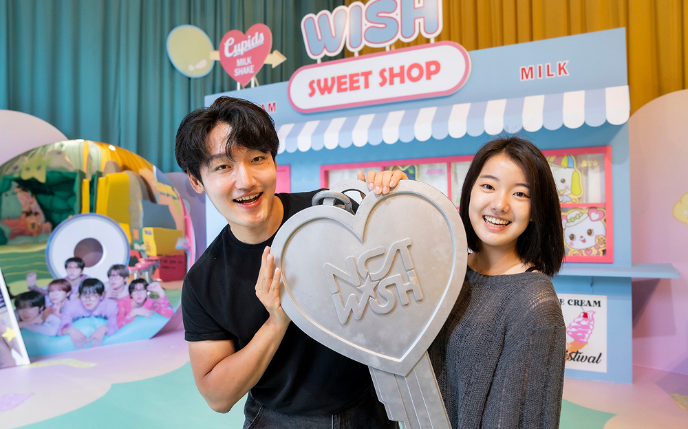 Two visitors smiling and holding a large heart-shaped "NCT WISH" key in front of a themed NCT WISH Sweet Shop, part of the EVER WISH collaboration at Everland Korea, operated by Samsung C&T Resort Group.