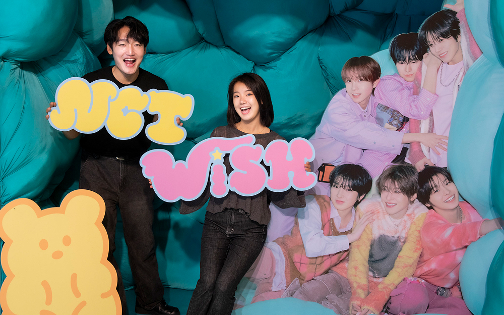Fans pose with an “NCT WISH” sign inside a themed photo zone at Everland Korea, featuring cutouts of the group members in a cozy, cloud-like setting.