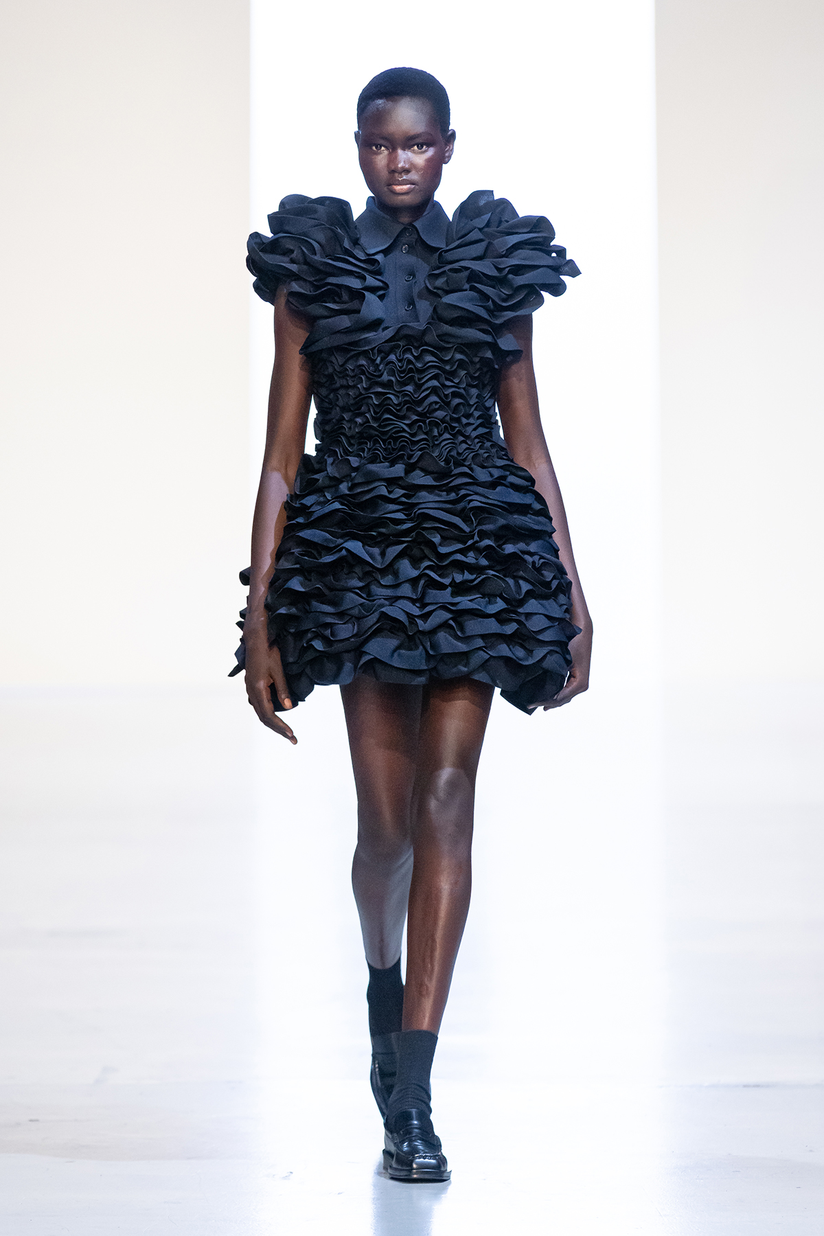 A model walks the runway in Juun.J’s 26SS collection wearing a voluminous black mini dress with dramatic layered ruffles and a structured collar, blending bold femininity with sculptural detail.