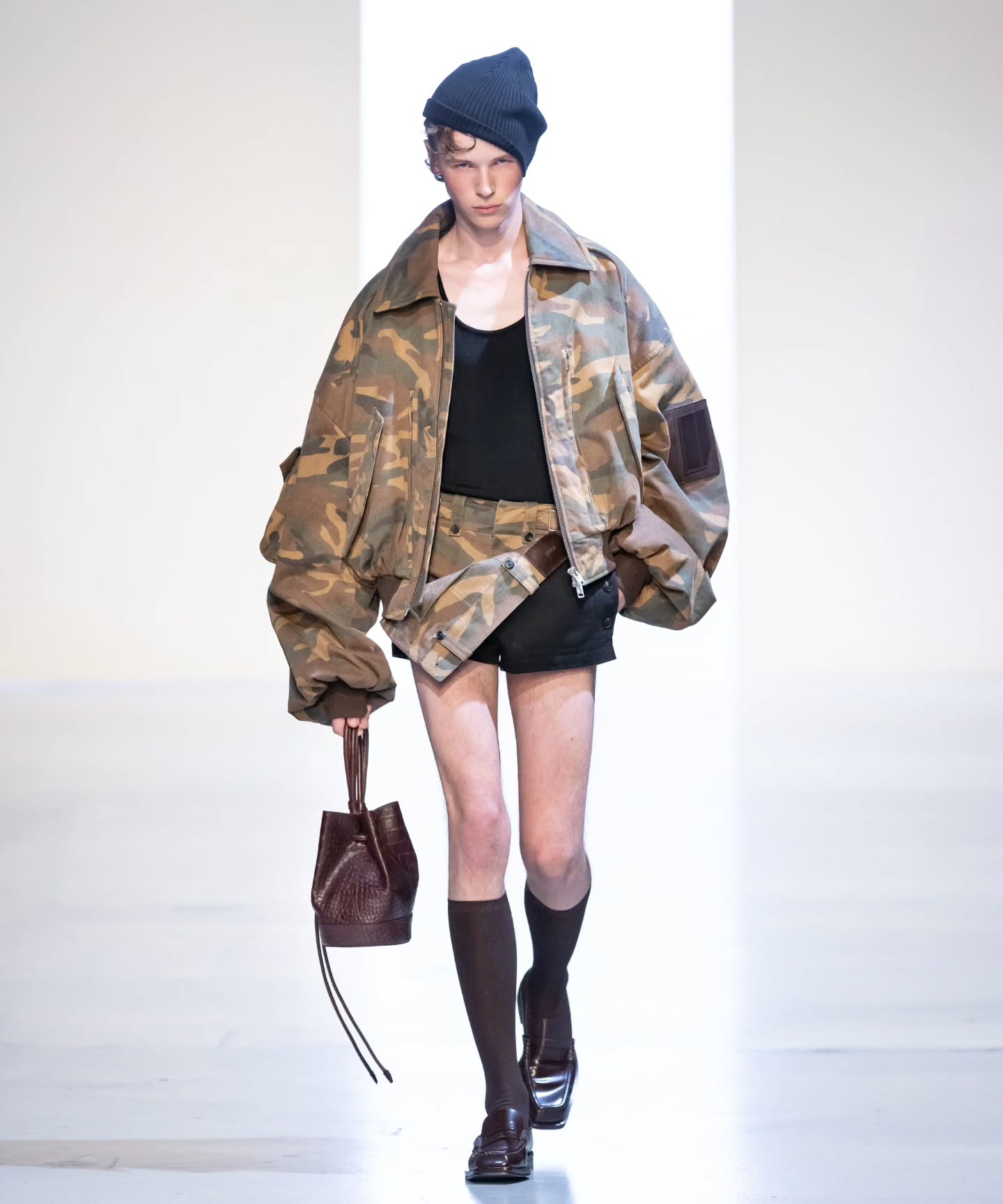 A model walks the runway in Juun.J’s 26SS collection wearing an oversized camouflage bomber jacket over a black tank top and micro mini skirt, styled with knee-high socks, loafers, and a brown leather bag for a bold streetwear look.
