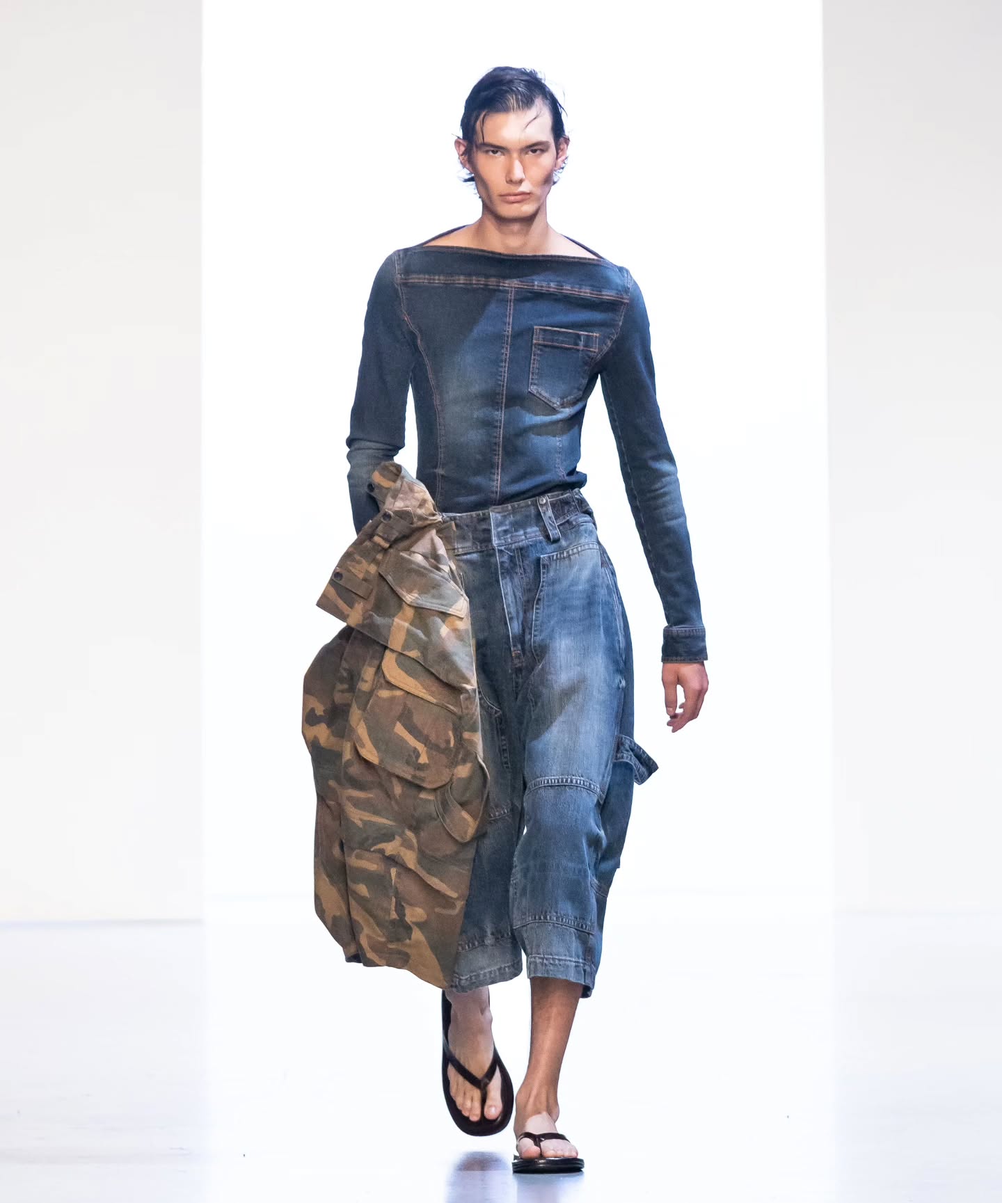 A model showcases Juun.J’s 26SS collection wearing a structured denim top with exposed stitching and wide cropped denim pants, styled with an oversized camouflage accent piece for a modern utilitarian aesthetic.