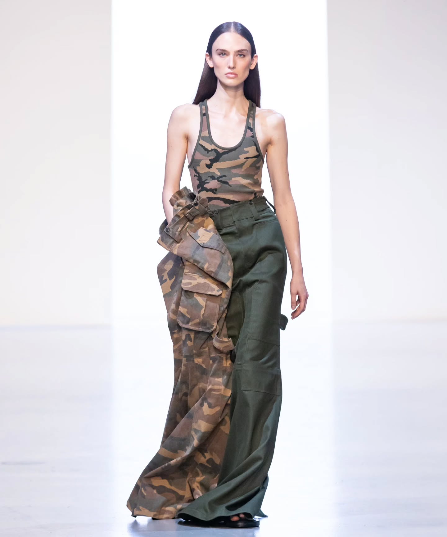 A model walks the runway in Juun.J’s 26SS collection wearing a fitted camouflage tank top and wide-leg green tailored trousers with an oversized camo fabric detail, creating a bold interplay of proportions and textures.