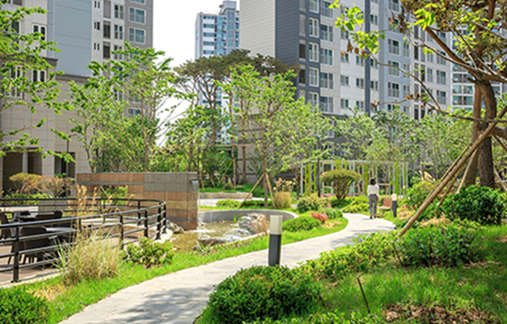 View of Raemian Artrich with landscaped gardens, wooden seating areas, walking paths, and surrounding apartment buildings. Trees and greenery create a pleasant residential environment.