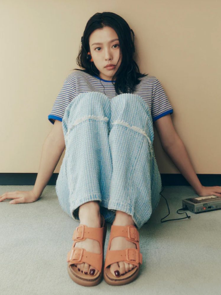 A woman poses in a pair of coral summer Scholl strappy sandals while seated on the floor, listening to an old school cassette player.