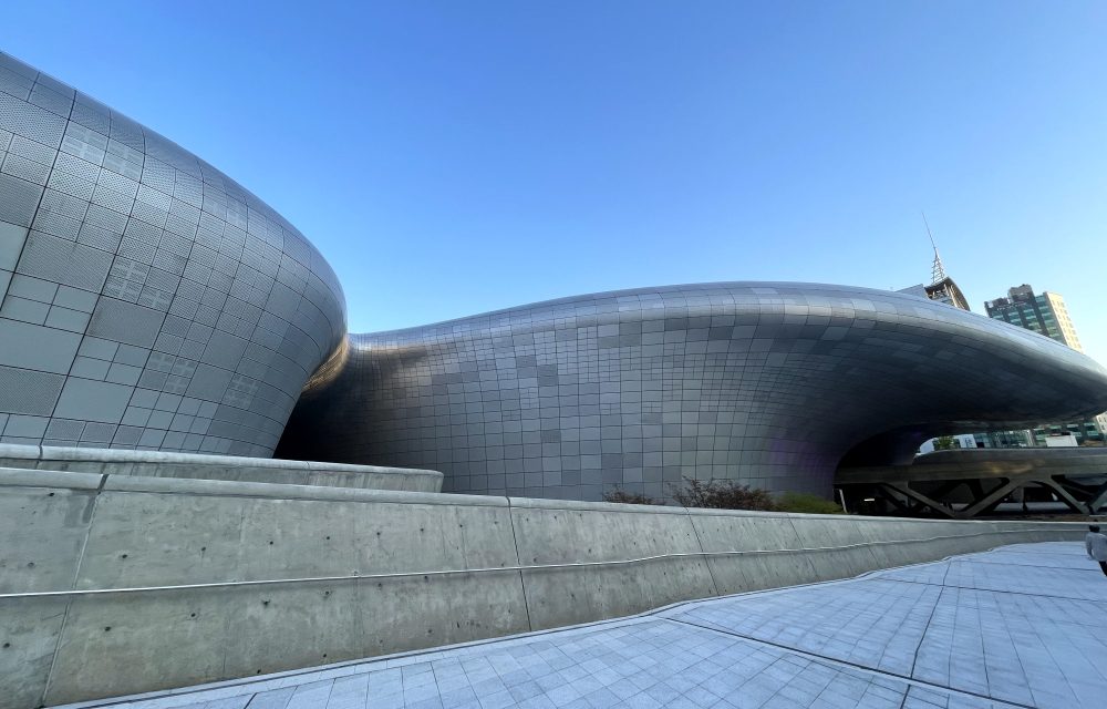 A low-angle view of the Dongdaemun Design Plaza (DDP) that highlights the smooth, curved surface of the building's aluminum panel facade, with a clear blue sky in the background.