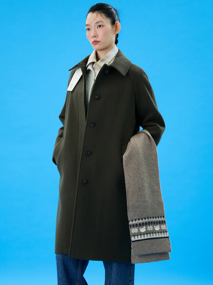 A model stands at a forty-five-degree angle in front of a bright blue wall and poses in an olive green tailored wool blend coat, paired with a collared knit jumper and a pair of slouchy jeans while she holds a long grey scarf that has Norwegian knitting patterns at each end.
