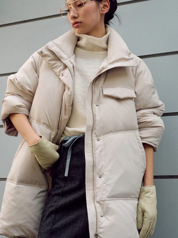 A woman poses against a wall in a long padded jacket in an ivory color, wearing gold aviator glasses.