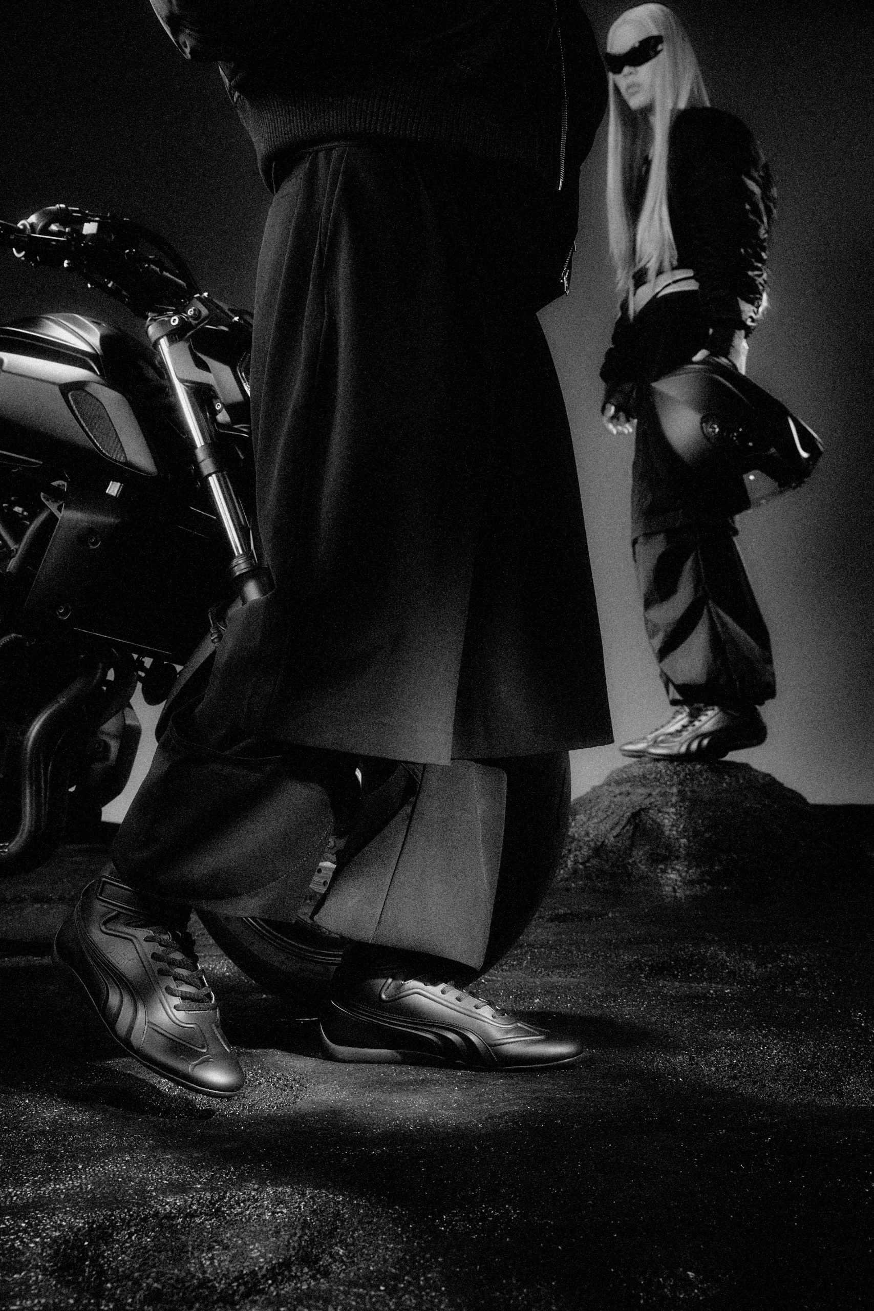 A model wearing Juun.J x PUMA Speedcat shoes stands beside a motorcycle, with another model in the background holding a helmet, both dressed in black with a modern, urban theme.
