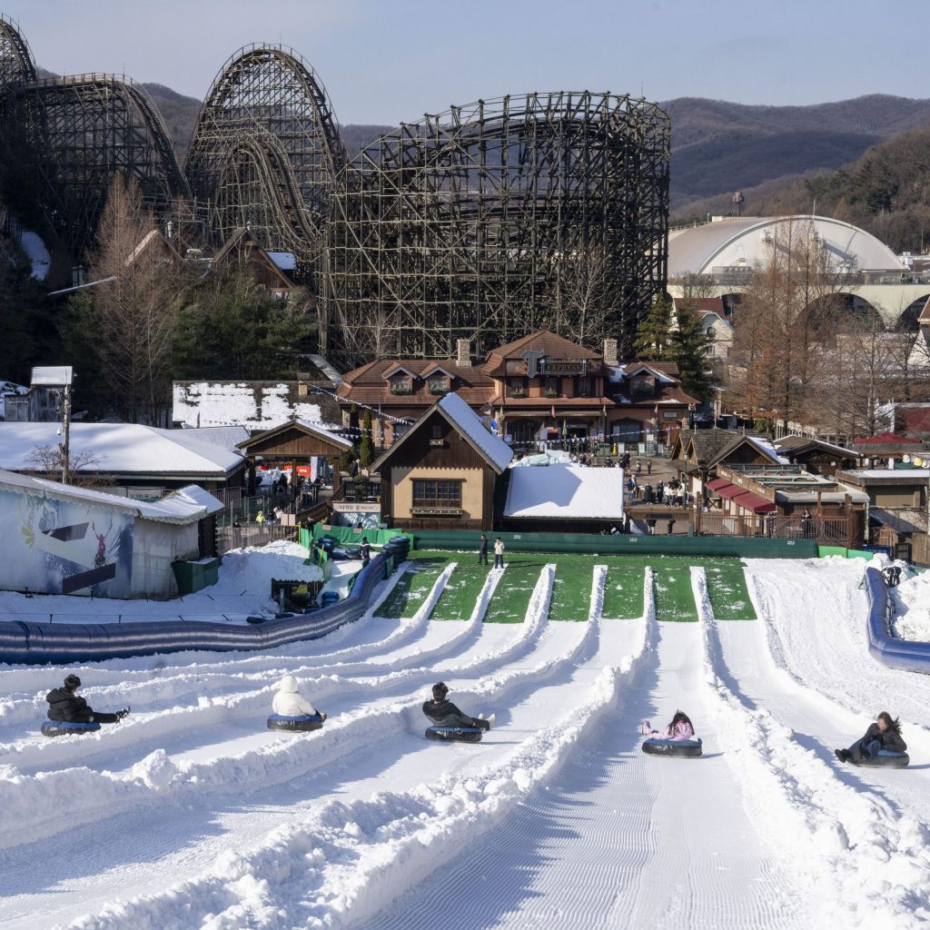Visitors sledding down multiple snowy lanes of the Racing Course at Everland, with a backdrop of wooden roller coasters and charming cottages under a bright winter sky.
