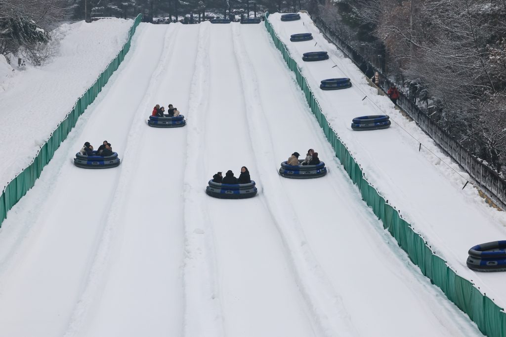 A wide-angle view of groups riding inflatable sleds down the Express Course at Everland, featuring long snowy tracks lined with green safety barriers under a snowy winter landscape.