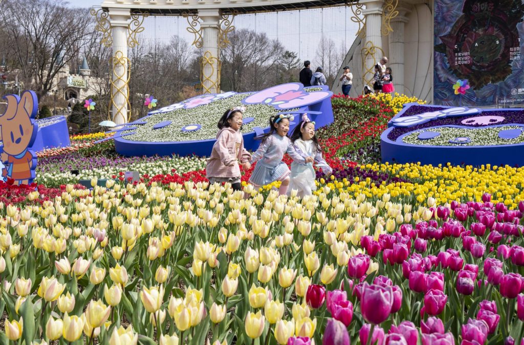 3 children hold hands and skip through a garden of colorful yellow and pink tulips at Everland Resort in South Korea. In the background, Sanrio characters Hello Kitty and Kuromi lie flat on a garden bed. A stage for outdoor performances is visible in the distance.