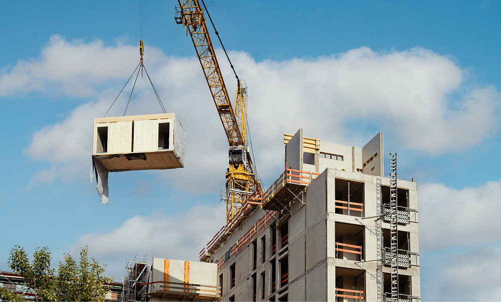 A construction crane lifts a prefabricated modular unit onto a partially completed building under a clear sky. The image illustrates Off-Site Construction (OSC) in action, where factory-built modules are assembled on-site to accelerate the construction process.