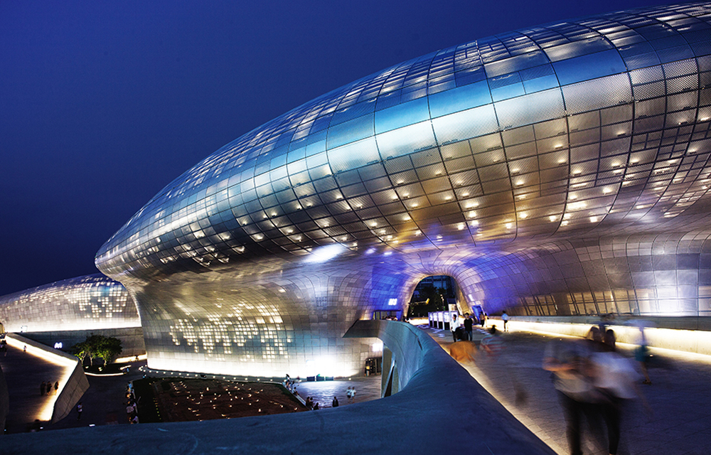 A nighttime view of Dongdaemun Design Plaza (DDP) in Seoul, South Korea, showcasing its futuristic curved architecture. The structure, built by Samsung C&T, is covered in 45,000 aluminum panels that were prefabricated off-site and installed on-site over a span of two years.