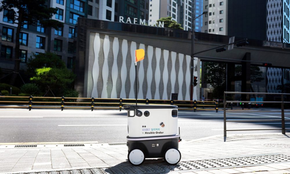 Deli-Pick Me delivery robot with a small orange flag is stationed on a pedestrian sidewalk in front of a Raemian apartment complex entrance.