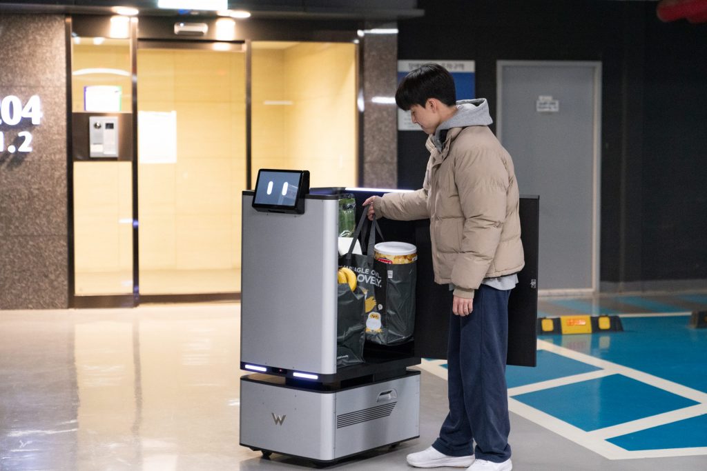 A young man in a beige jacket removes groceries from a Carry Robot in an underground parking facility. The robot’s digital face display and secure storage compartment highlight its role in automated residential convenience.