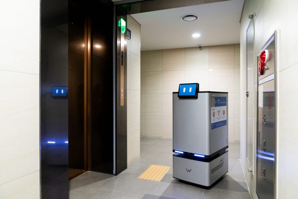 A Carry Robot in a well-lit apartment corridor positioned in front of an elevator, ready to autonomously board and navigate to a resident’s floor. The robot’s sleek design and blue-lit display contrast with the neutral-colored walls and flooring.
