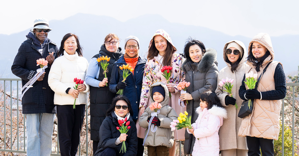 A group of adults and children from various diplomatic families smile while holding colorful tulip bouquets at Everland, with mountains in the background.