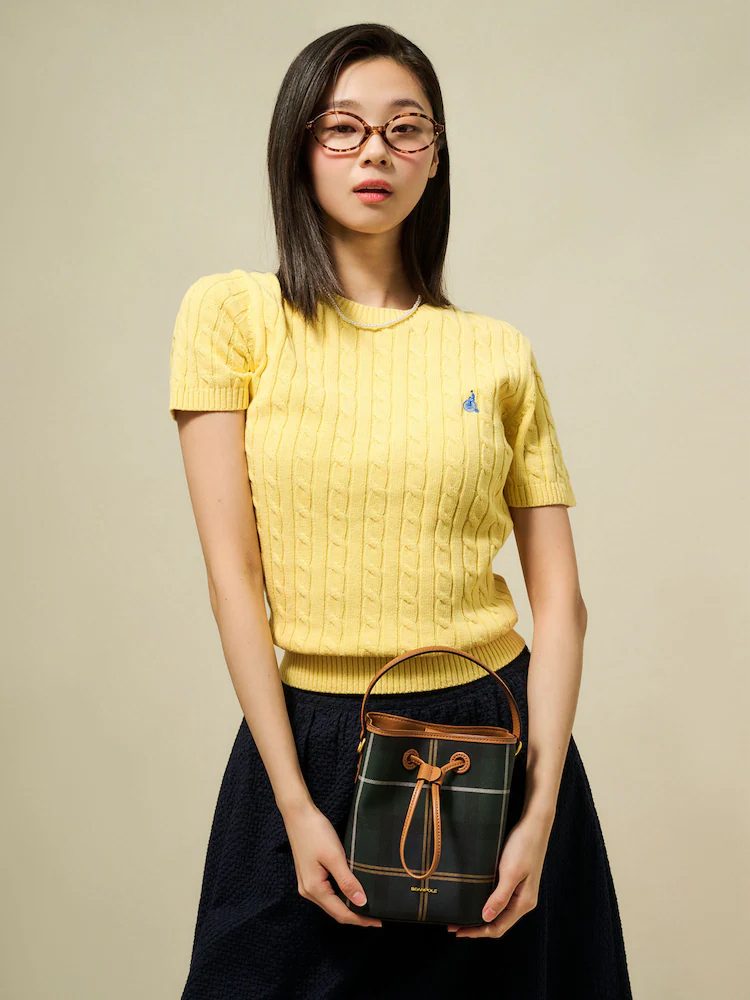 A model wears the Cable Round Neck Short-Sleeve Pullover in yellow from Beanpole’s SolSol Knit collection. Paired with a navy textured skirt and a green check bucket bag with leather accents, the look balances youthful energy with timeless elegance.
