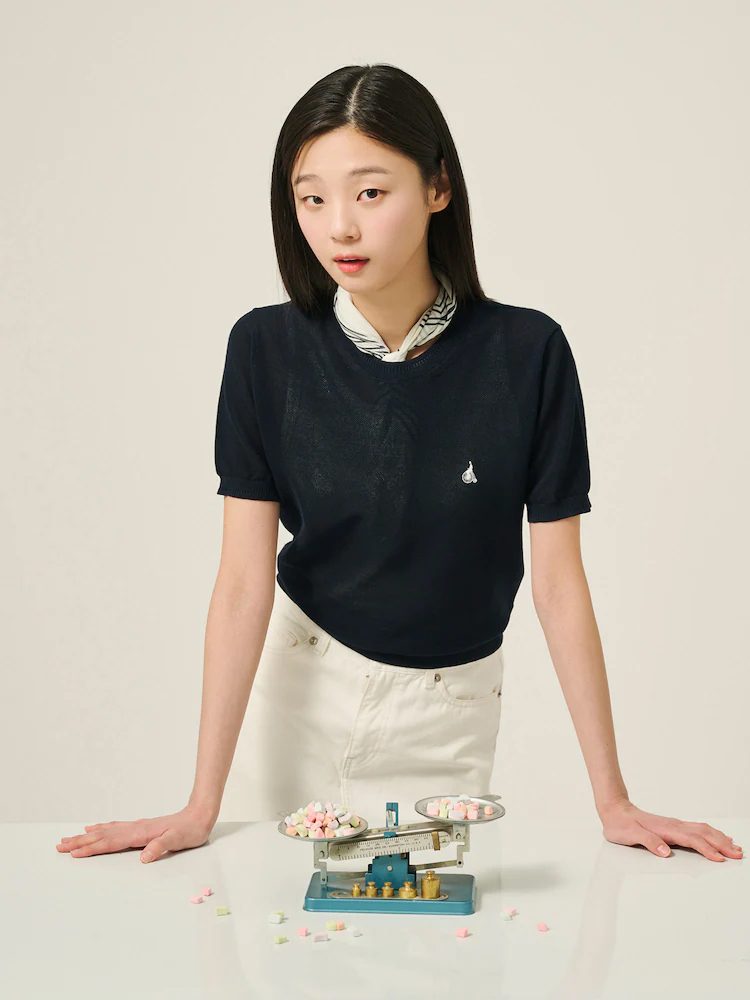 A female model wears Beanpole’s Honeycomb Round Neck Short-Sleeve Knit in navy, styled with a printed scarf and white pants, standing behind a table with a vintage blue balance scale holding pastel candies for a playful yet minimalist spring look