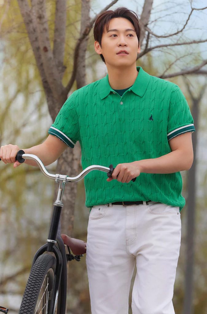 John Park wears the Cable Collar Neck Knit in apple green from Beanpole’s SolSol Knit collection. Styled with white jeans and layered subtly over a darker tee, the look is captured as he walks a bike through a serene, tree-lined setting, reflecting casual spring freshness.