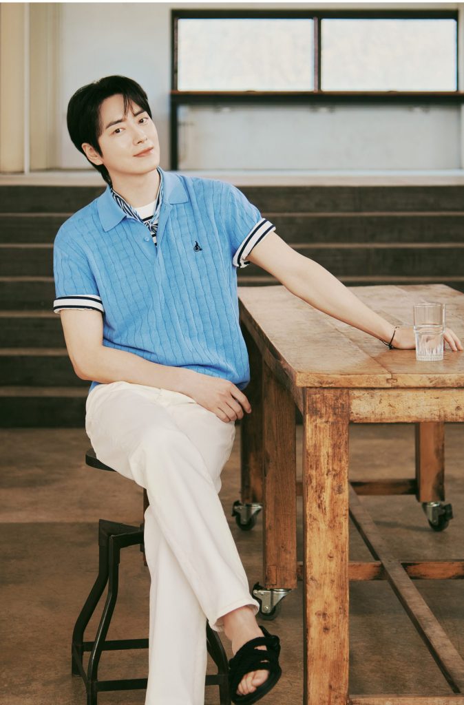 Lee Jun-hyuk is seated at a wooden table, wearing the Cable Collar Neck Knit in sky blue from Beanpole’s SolSol Knit collection. The relaxed setting and striped detailing reflect the collection’s refined and breathable spring aesthetic.