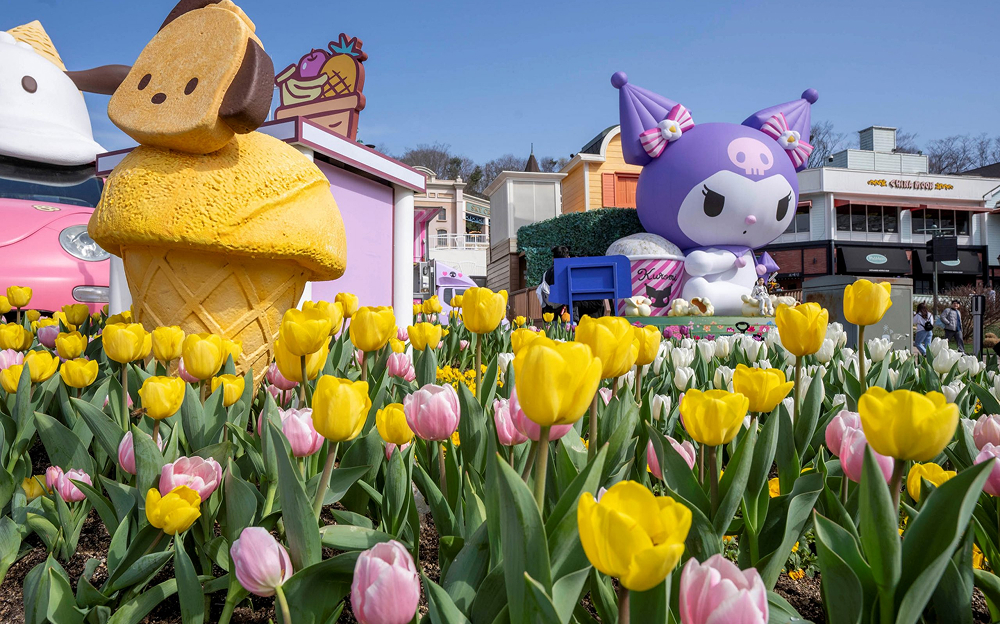 A colorful tulip garden with yellow, pink, and white tulips in full bloom. Giant statues of Sanrio characters, including Pompompurin in an ice cream cone and Kuromi, decorate the flower beds under a clear blue sky.