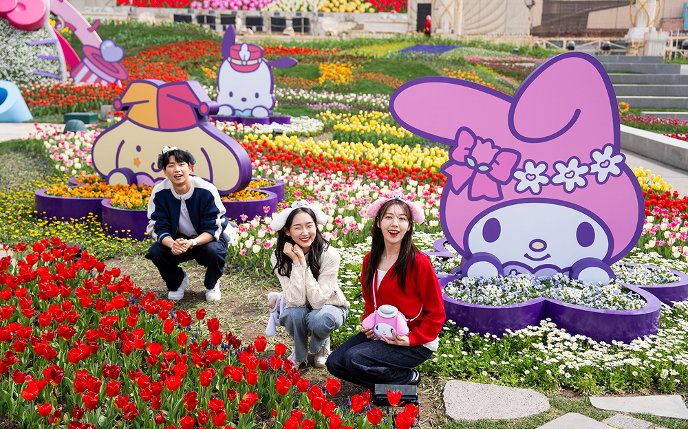 Three young adults pose cheerfully among blooming red tulips, surrounded by large My Melody, Cinnamoroll, and Pompompurin cutouts in a brightly colored floral display.