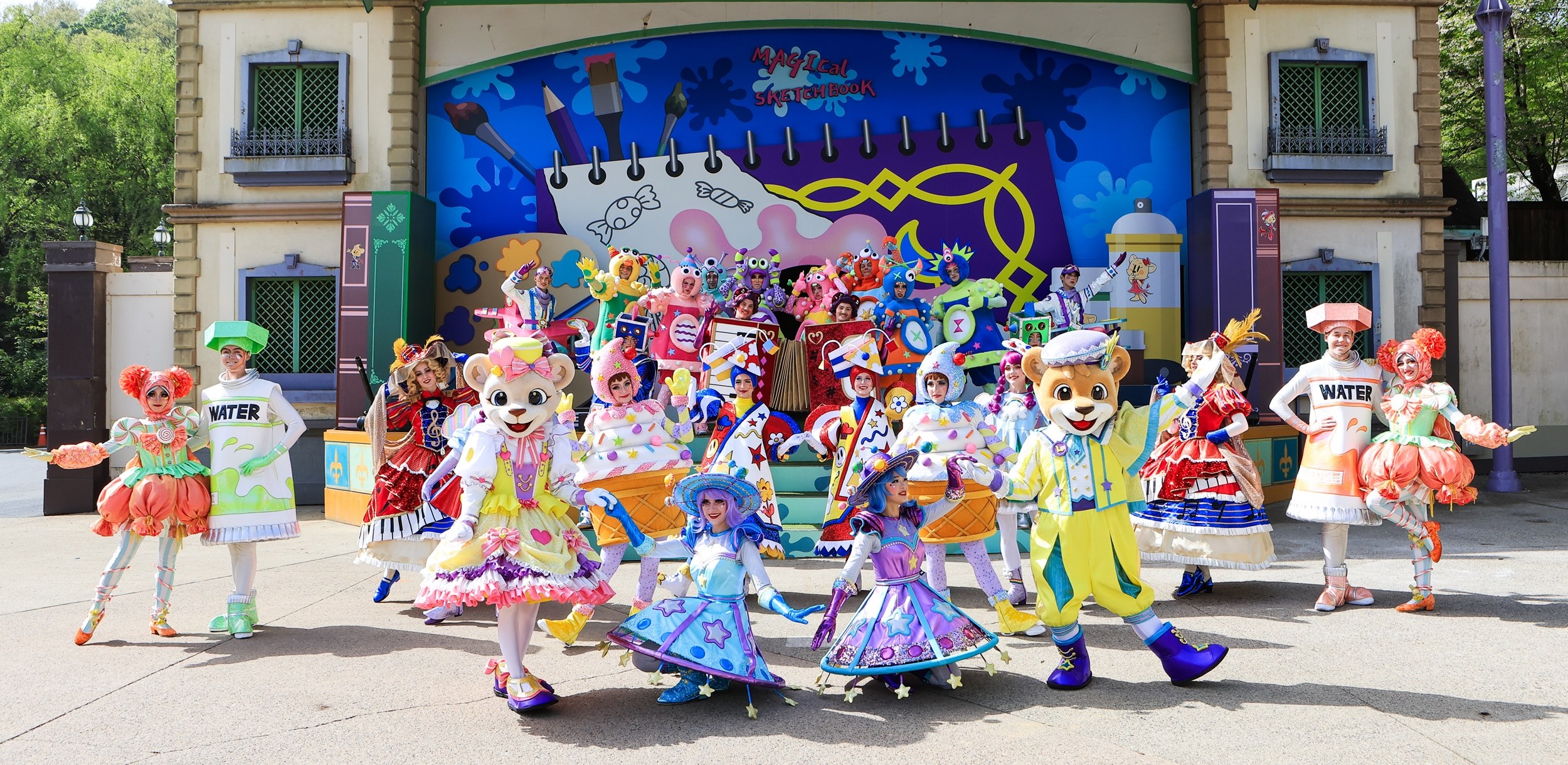 A group of performers in vibrant, themed costumes pose in front of the Magical Sketchbook stage at Everland, with mascots Lenny and Lara leading a cheerful ensemble.