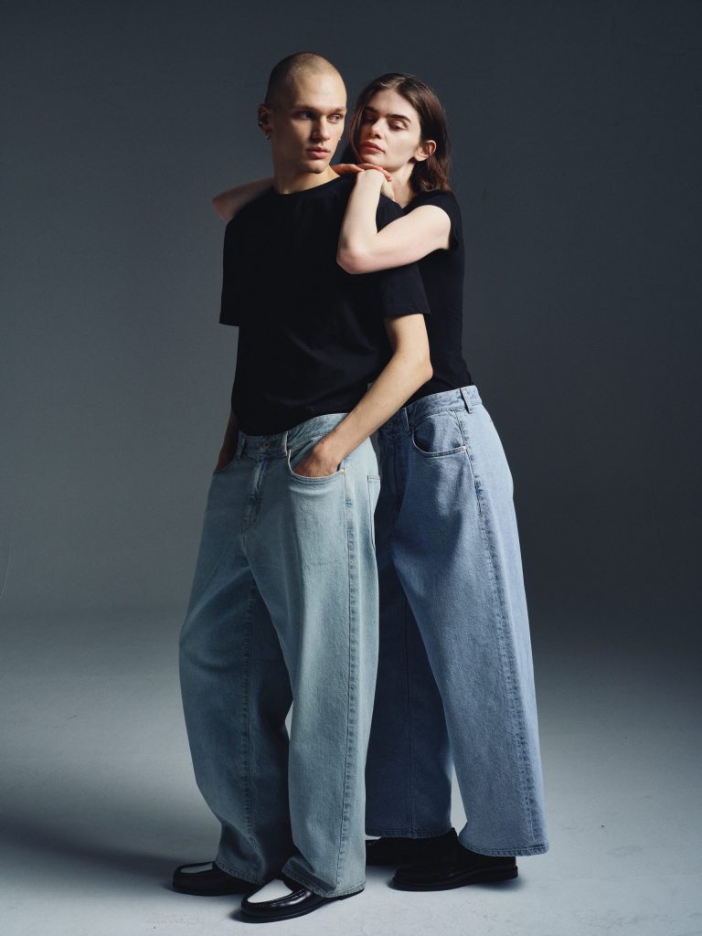 Two models wear black T-shirts with oversized light-wash denim jeans. Their intertwined pose and tonal palette express ease, comfort, and effortless coordination.