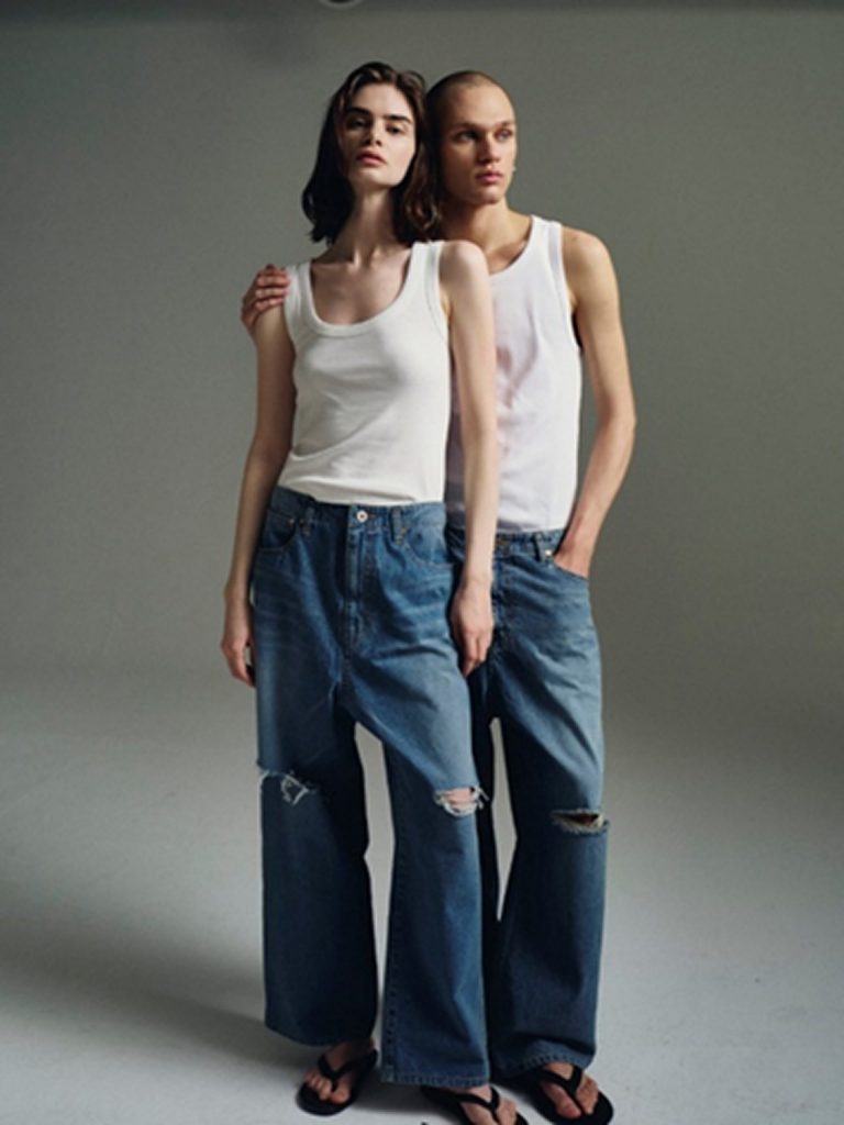 A male and female model stand close together in a studio, both wearing white tank tops and relaxed-fit blue jeans. The woman’s jeans feature distressed knees, while the man’s have a cleaner wash, highlighting contrast and connection.