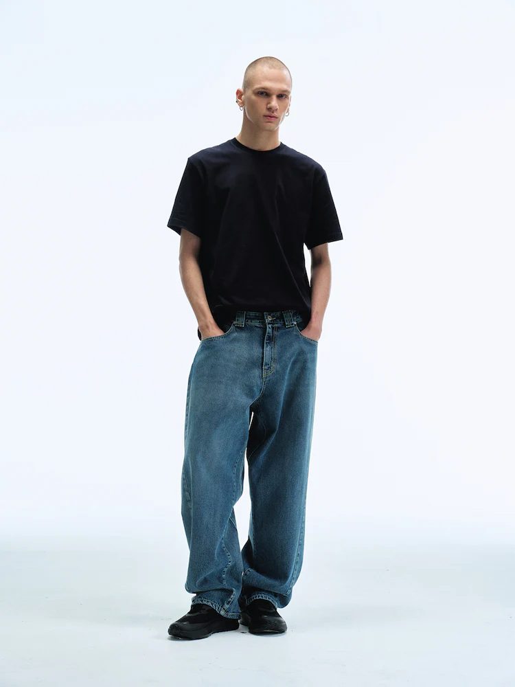 A male model poses in a studio wearing Men Reworked Wide Fit Denim Pants in Light Blue with a black short-sleeve T-shirt and black shoes, highlighting the pants’ relaxed volume and reworked design details.