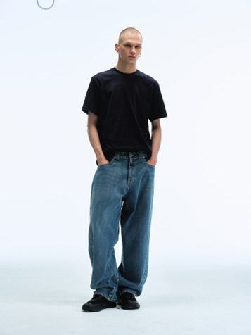 A male model poses in a studio wearing Men Reworked Wide Fit Denim Pants in Light Blue with a black short-sleeve T-shirt and black shoes, highlighting the pants’ relaxed volume and reworked design details.