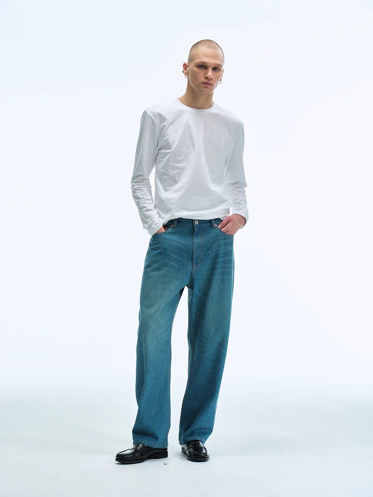 A male model stands in a studio wearing Men Straight Fit Denim Pants in Blue with a white long-sleeve T-shirt and black shoes, showcasing a relaxed yet refined silhouette.