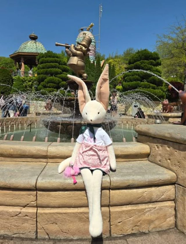 A plush bunny character dressed in a pink skirt and white Rrt t-shirt sits in front of a whimsical fountain at Everland, with a brass statue and lush greenery in the background.