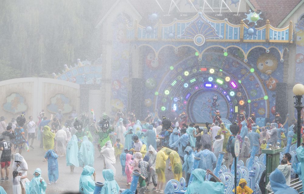 Families and children in colorful rain ponchos play in front of a decorative stage with bright LED lights and cartoon designs. Water sprays from above as the crowd gets soaked during the performance.