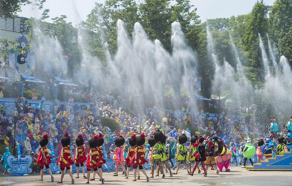 A colorful outdoor performance at Everland featuring dancers in vibrant costumes and oversized character heads. Behind them, fountains shoot water high into the air as crowds in ponchos watch from the stands.