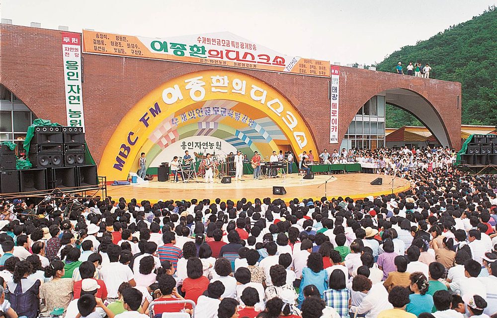 A large audience fills the outdoor venue during a daytime broadcast of “Lee Jong-hwan’s Disco Show” at Everland’s Rose Garden, with musicians and hosts performing on a bright stage framed by bold typography and curved architecture.