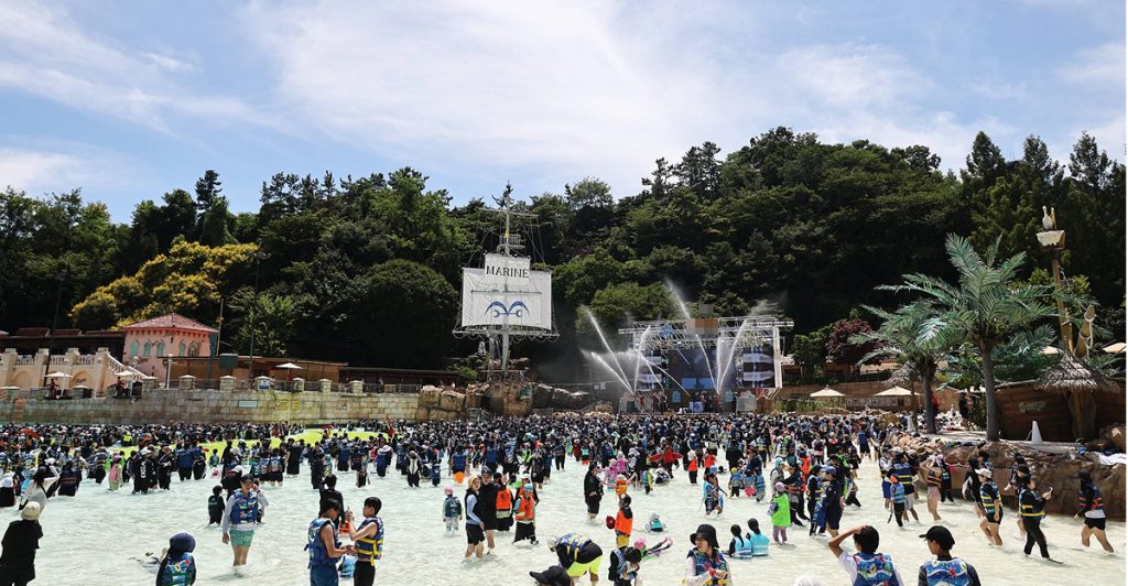 A wide crowd of visitors plays in the Caribbean Bay wave pool under a bright summer sky, with a pirate ship stage and water fountains in the background.
