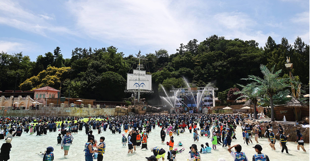 A wide crowd of visitors plays in the Caribbean Bay wave pool under a bright summer sky, with a pirate ship stage and water fountains in the background.