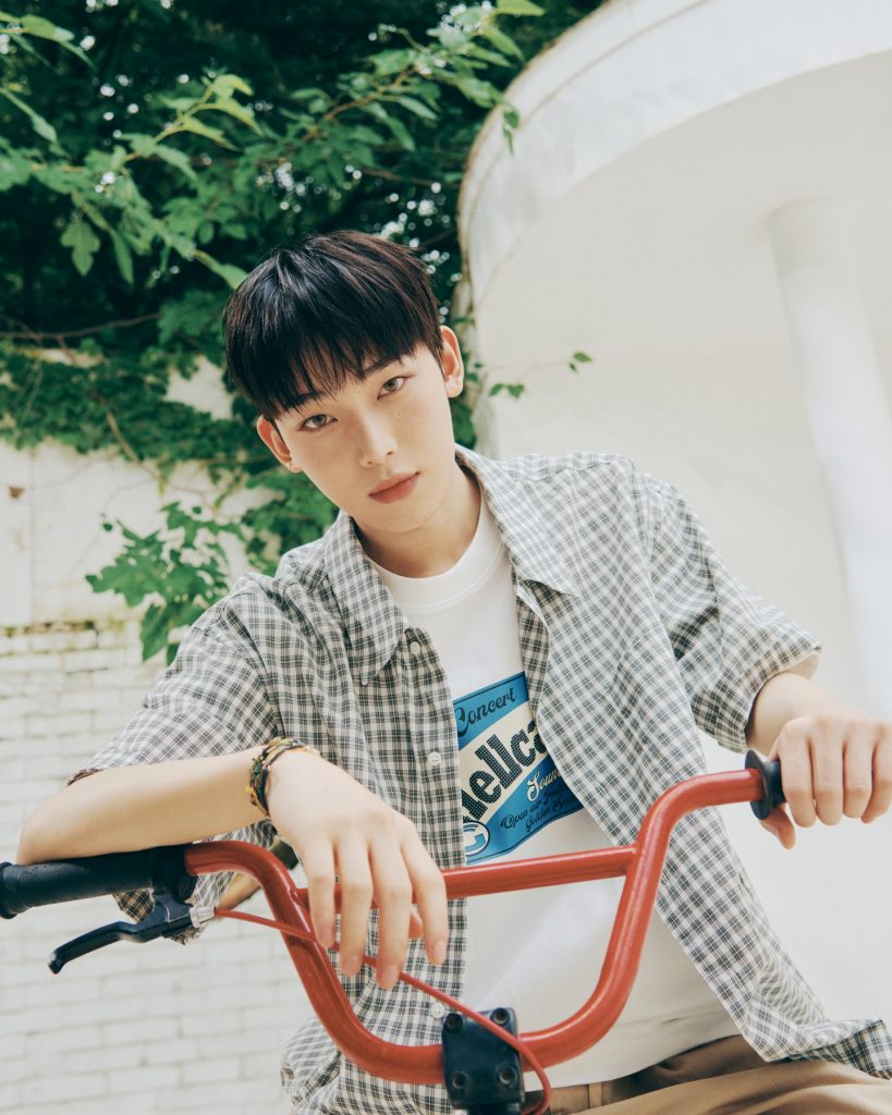 Sunoo leans forward on the handlebars of a red bicycle, wearing a checkered short-sleeve shirt over a white concert-print T-shirt. With greenery in the background, the photo evokes a retro, summery atmosphere.