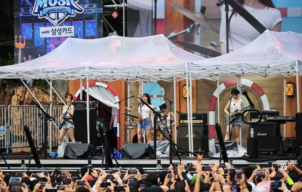 The girl group QWER performs on stage at Caribbean Bay during the Water Music Pool Party, with fans raising phones to capture the moment.