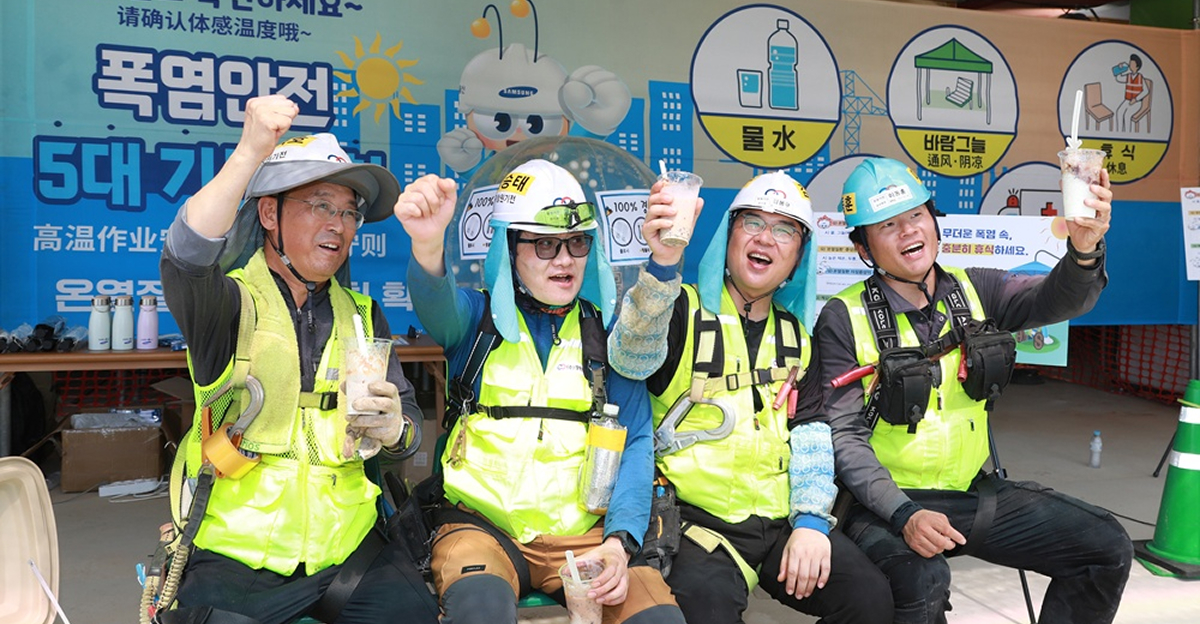 Four construction workers at a Samsung C&T site raise iced drinks and smile in front of a heat safety banner encouraging hydration and cooling.