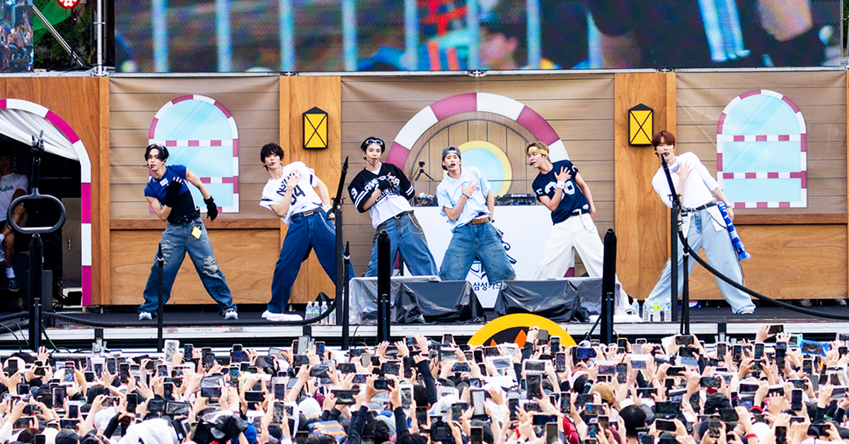 K-pop group RIIZE performs in coordinated summer outfits on an outdoor stage at Caribbean Bay, as a large crowd watches and records with phones raised.