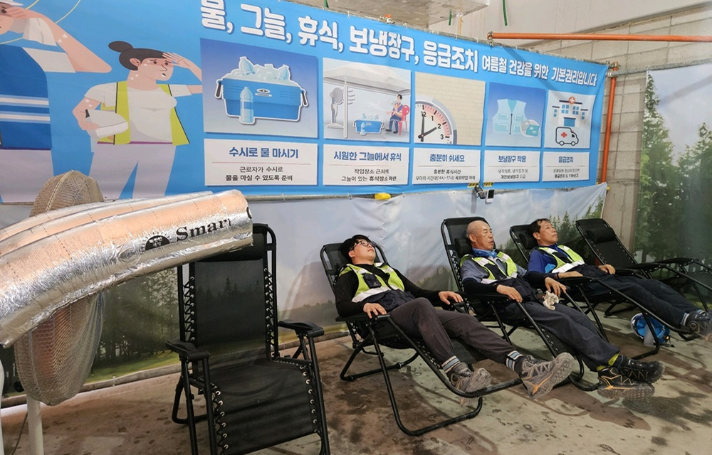 Three construction workers resting in reclining chairs under a cooling fan inside a Samsung C&T site, in front of a safety poster about hydration and rest.