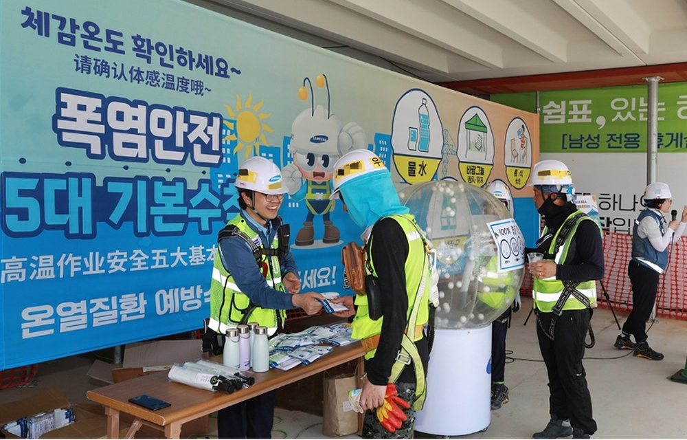 Construction workers in safety vests receiving cooling supplies and information at a Samsung C&T heat safety campaign booth with posters and banners.