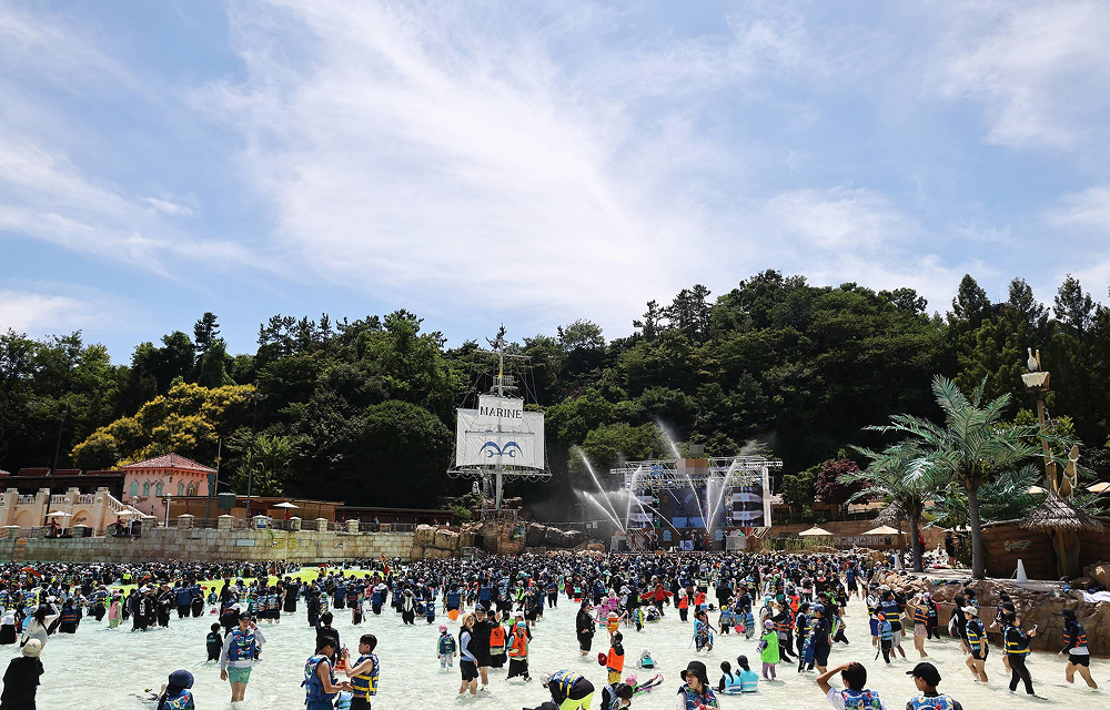 A wide view of Caribbean Bay’s wave pool filled with guests in swimsuits and life vests, with a pirate ship stage and forest backdrop under a clear summer sky