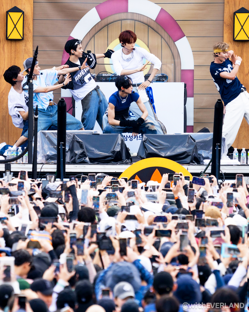 RIIZE performs choreographed moves on stage in front of a large crowd at Caribbean Bay, with hundreds of fans holding up phones to record the performance.