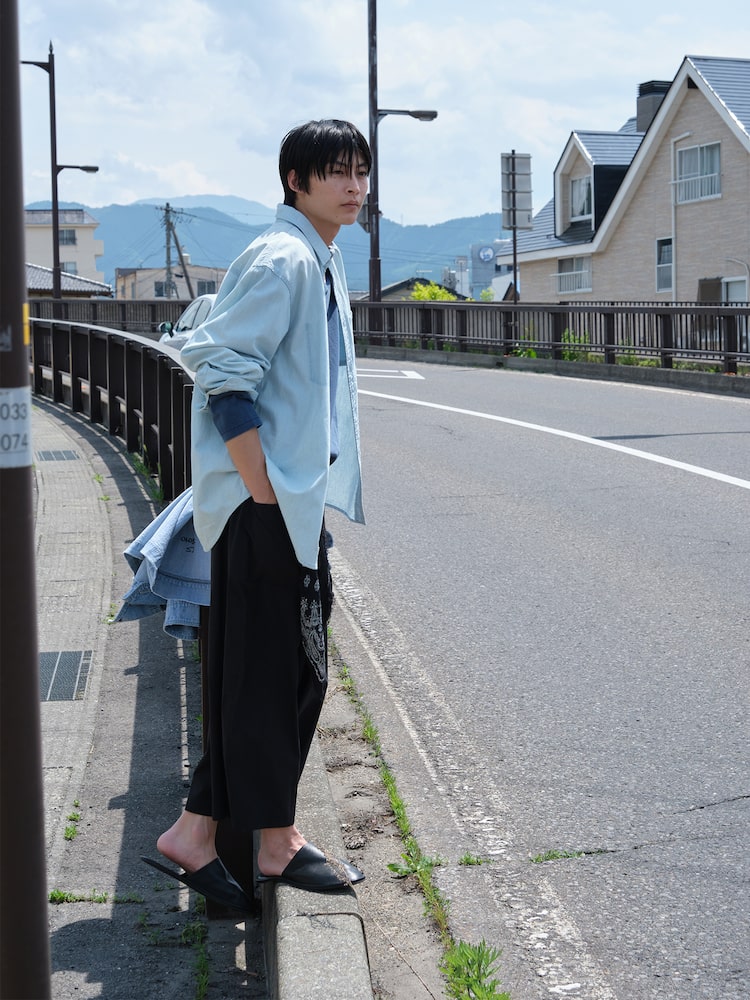 Model standing on a roadside wearing the Beaker Original Men Striped Denim Shirt in Sky Blue, styled loosely over black wide-leg trousers and slip-on shoes. The shirt is made from 100% cotton denim and designed for an easy oversized silhouette.