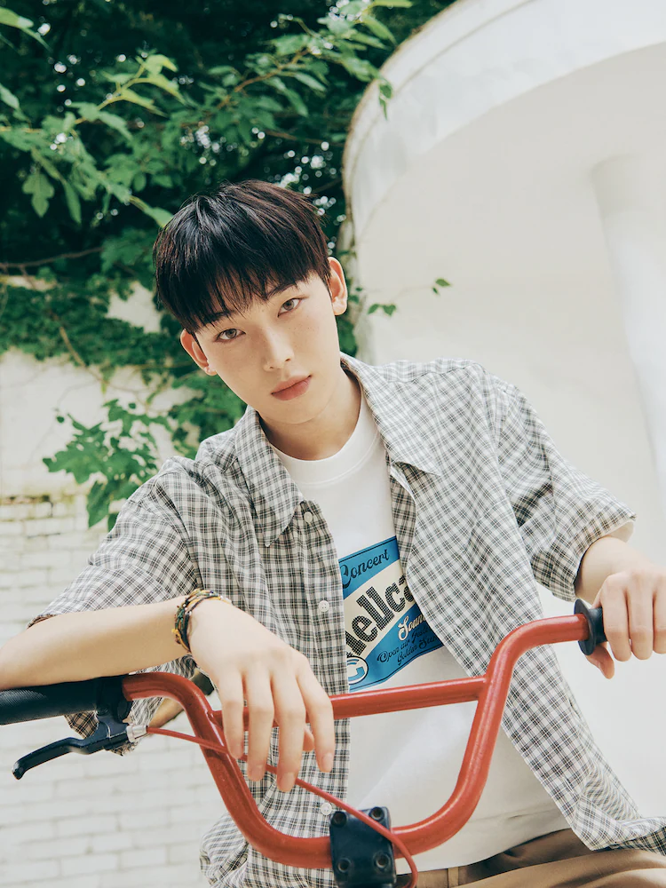 Male model outdoors wearing the 8seconds Short-Sleeve Check Shirt in Green, styled open over a graphic T-shirt while leaning on a bicycle. The shirt is made from 100% cotton and features a relaxed, oversized fit designed for breathable comfort.